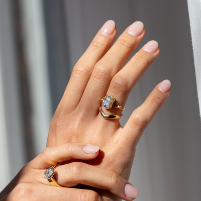 Hand wearing two gold rings with gemstones against a neutral background