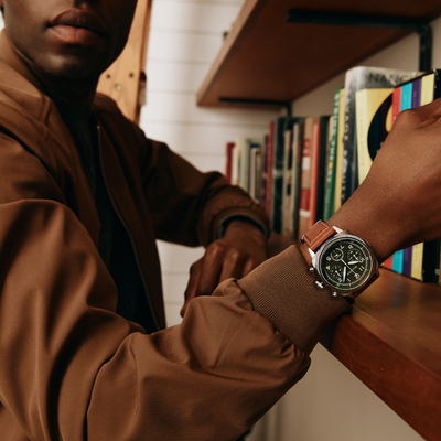 A close-up lifestyle shot of a man wearing a silver-toned chronograph watch with a brown leather strap, leaning his arm on a wooden bookshelf.