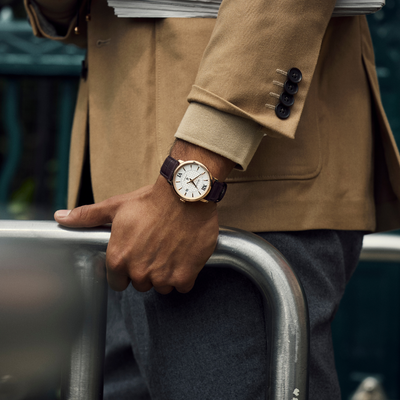 Person wearing a brown coat and holding a newspaper, with a close-up of a wristwatch.  A Gent's Bulova Brown Strap Hudson Automatic Watch.
