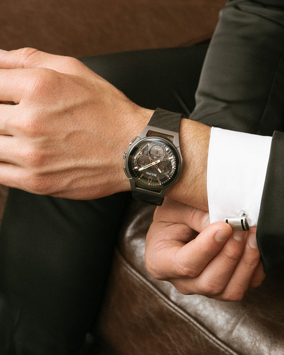 Close-up of a person wearing a dark wristwatch with a detailed face, sitting on a brown leather couch.