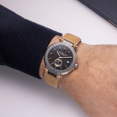 A wristwatch with a black dial, silver-toned case, and tan leather strap worn on a person's wrist, with a dark ribbed sweater sleeve visible.