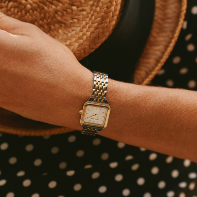 A close-up of a person's wrist wearing a gold and silver-toned rectangular watch with a white dial, styled with a woven straw hat and a black dress with white polka dots.
