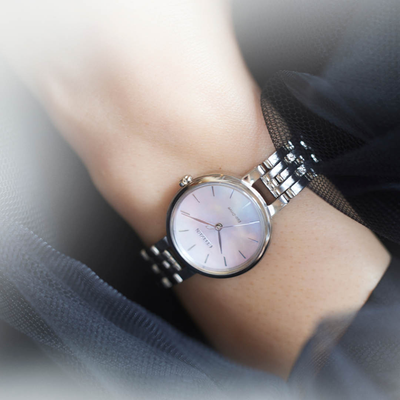 A close-up of a woman's wrist wearing a minimalist analog watch with a silver metal link bracelet, a round silver case, and a pale pink dial. The brand name 'SOLIMA' is visible on the watch face.