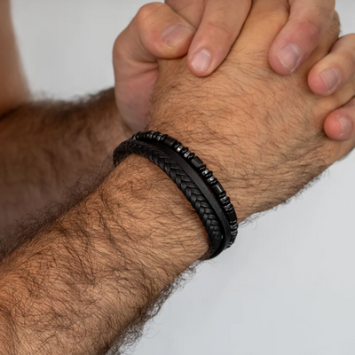 A close-up of a man's wrist wearing two black bracelets, one braided and one beaded, against a light gray background.