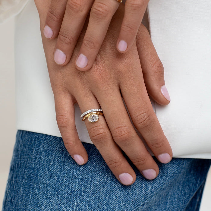 Close-up of a person's hands wearing multiple rings, including a yellow gold band with a heart-shaped clear gemstone and a silver-toned band with a round clear gemstone, styled with a white top and blue jeans.