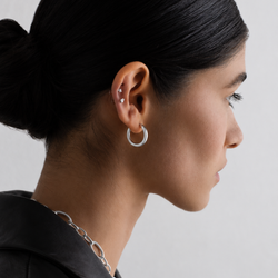 A close-up side profile of a woman with dark hair in a bun, wearing a polished silver hoop earring and a silver chain necklace against a light gray background.