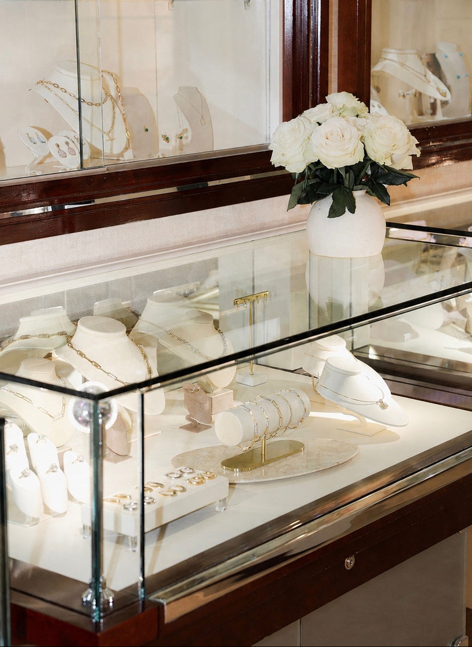 A glass display case in a jewelry store showcasing white jewelry stands, necklaces, rings, and earrings, with a vase of white roses on top.