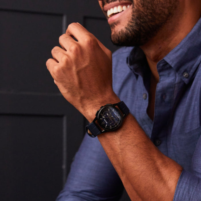 Man wearing a blue shirt and black watch, smiling outdoors.